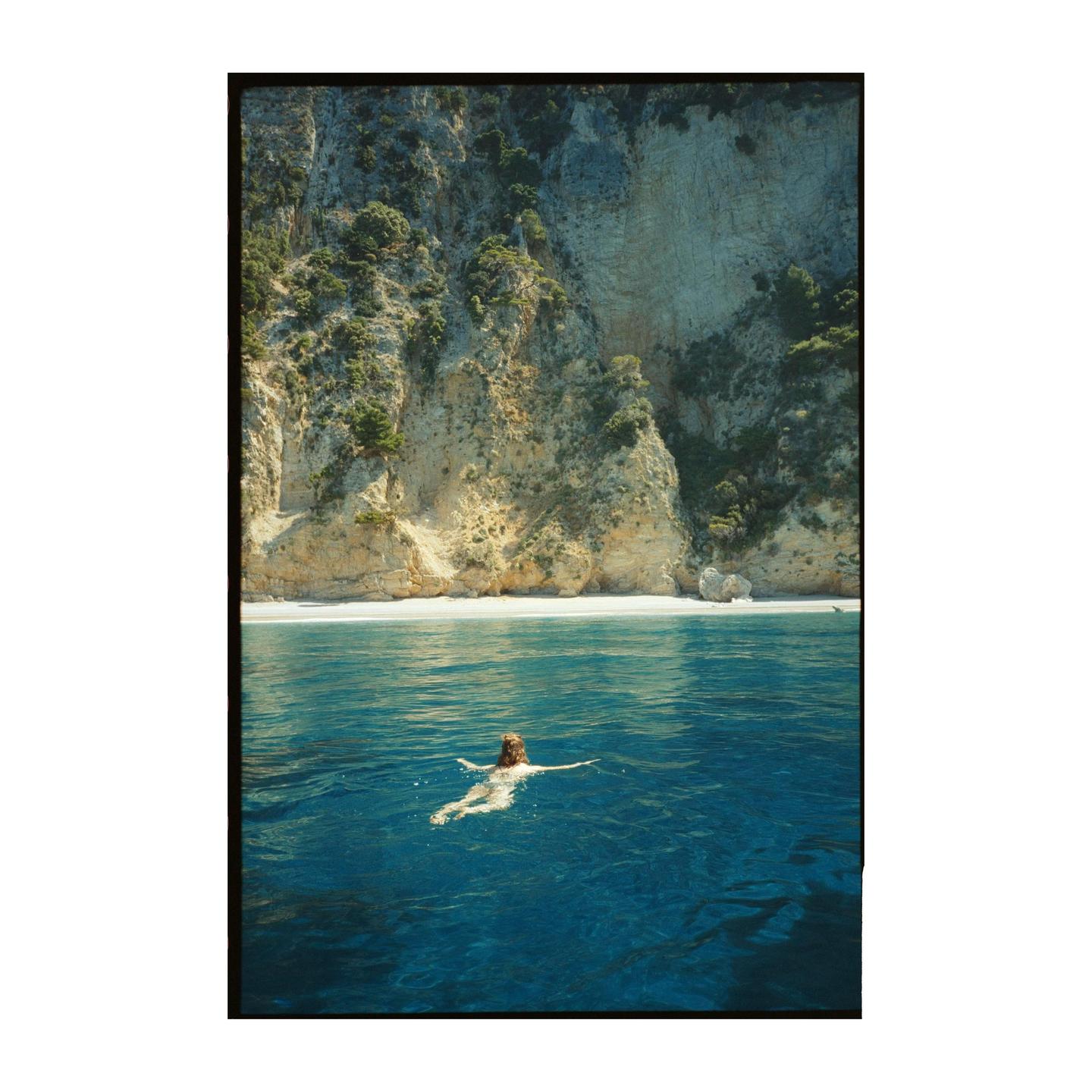 A woman swims in clear blue water near a white sand beach backed by a large cliff.