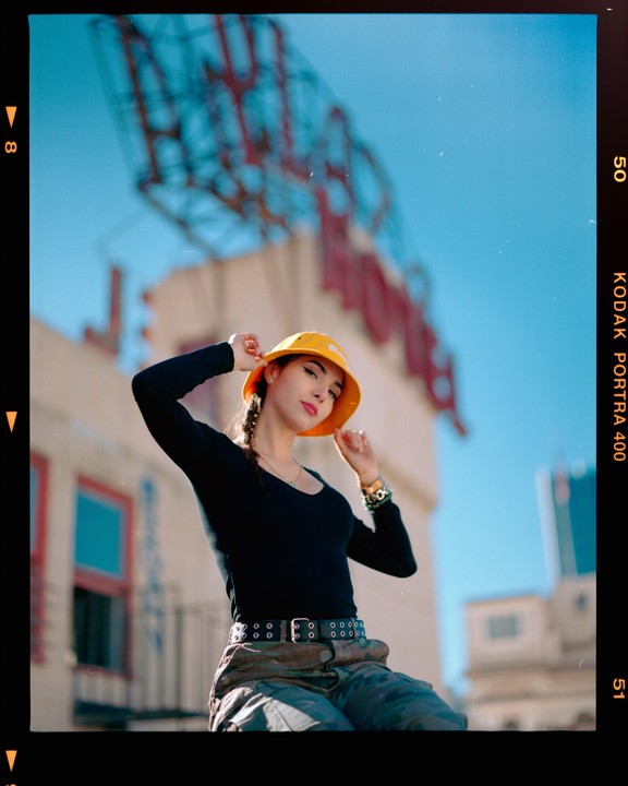 A woman in a yellow bucket hat and black shirt poses outdoors with a building and blue sky in the background.