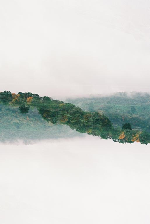 A double exposure shows a forest with autumn foliage reflected in a body of water, with fog in the background.