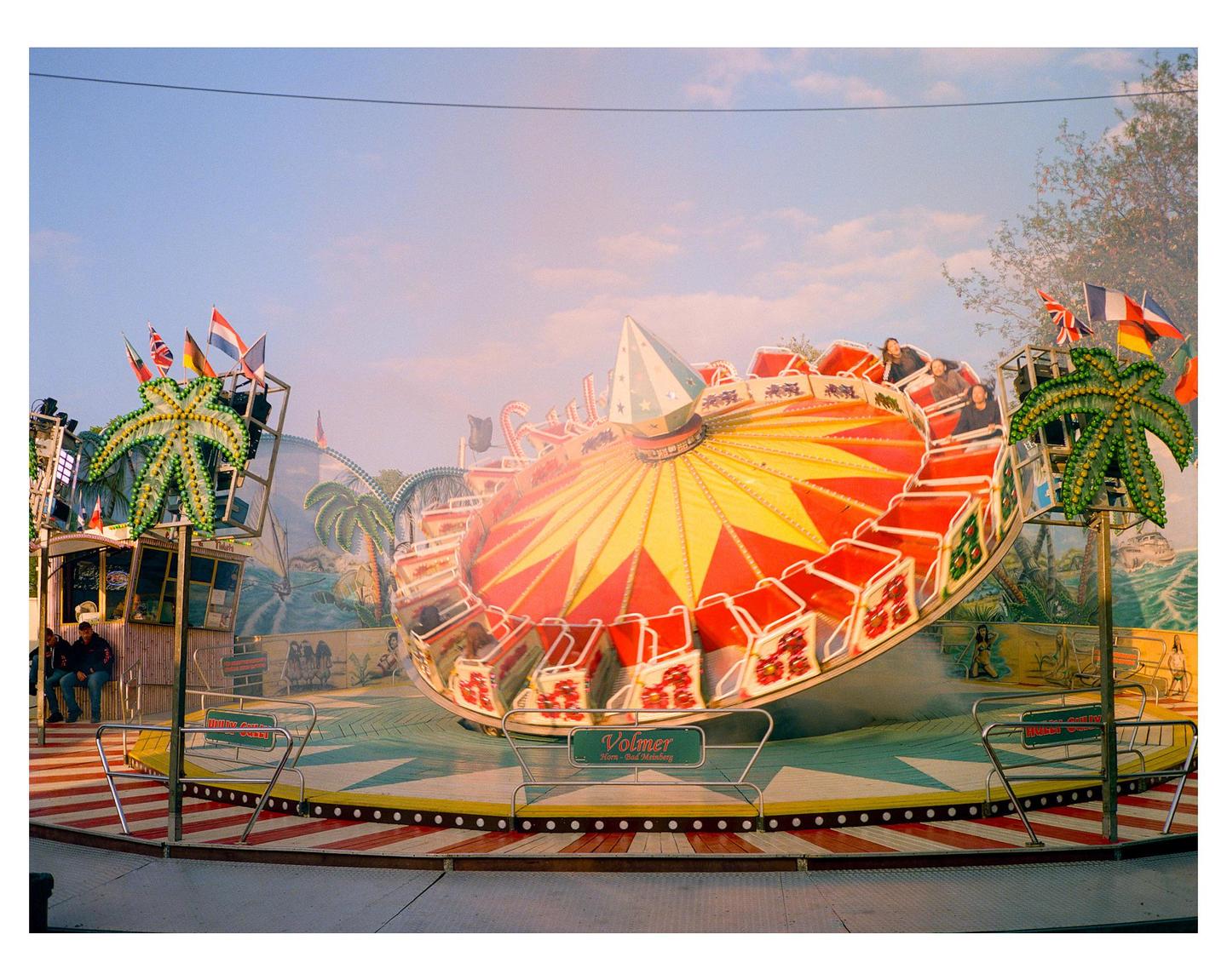 A spinning carnival ride with people on it is surrounded by palm tree decorations and flags under a clear sky.