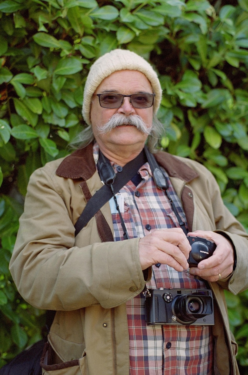 A man with a mustache and glasses wears a beanie and a plaid shirt while holding a camera.
