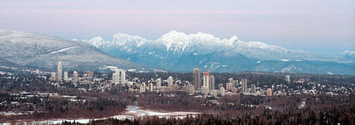 A city with tall buildings is nestled among snow-covered trees and mountains under a pale sky.