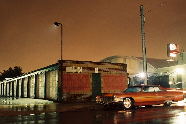 A classic car is parked on a wet street next to a row of garages and a brick building with a "RENT" sign at night.