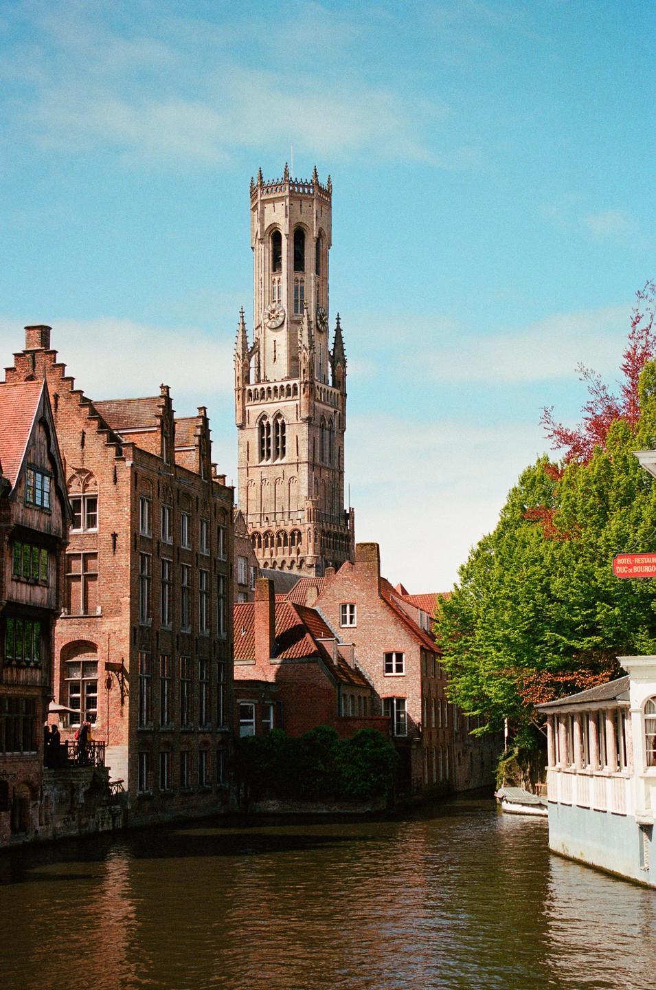 A tall, ornate bell tower rises above brick buildings and a canal.