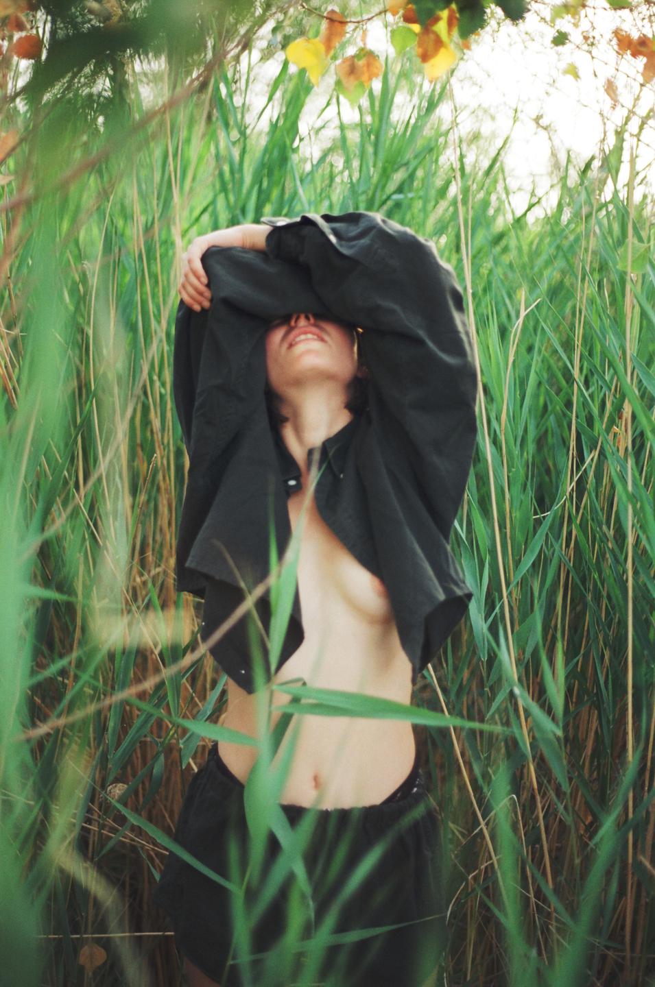 A woman stands in tall grass with her shirt pulled over her head, revealing her bare chest.