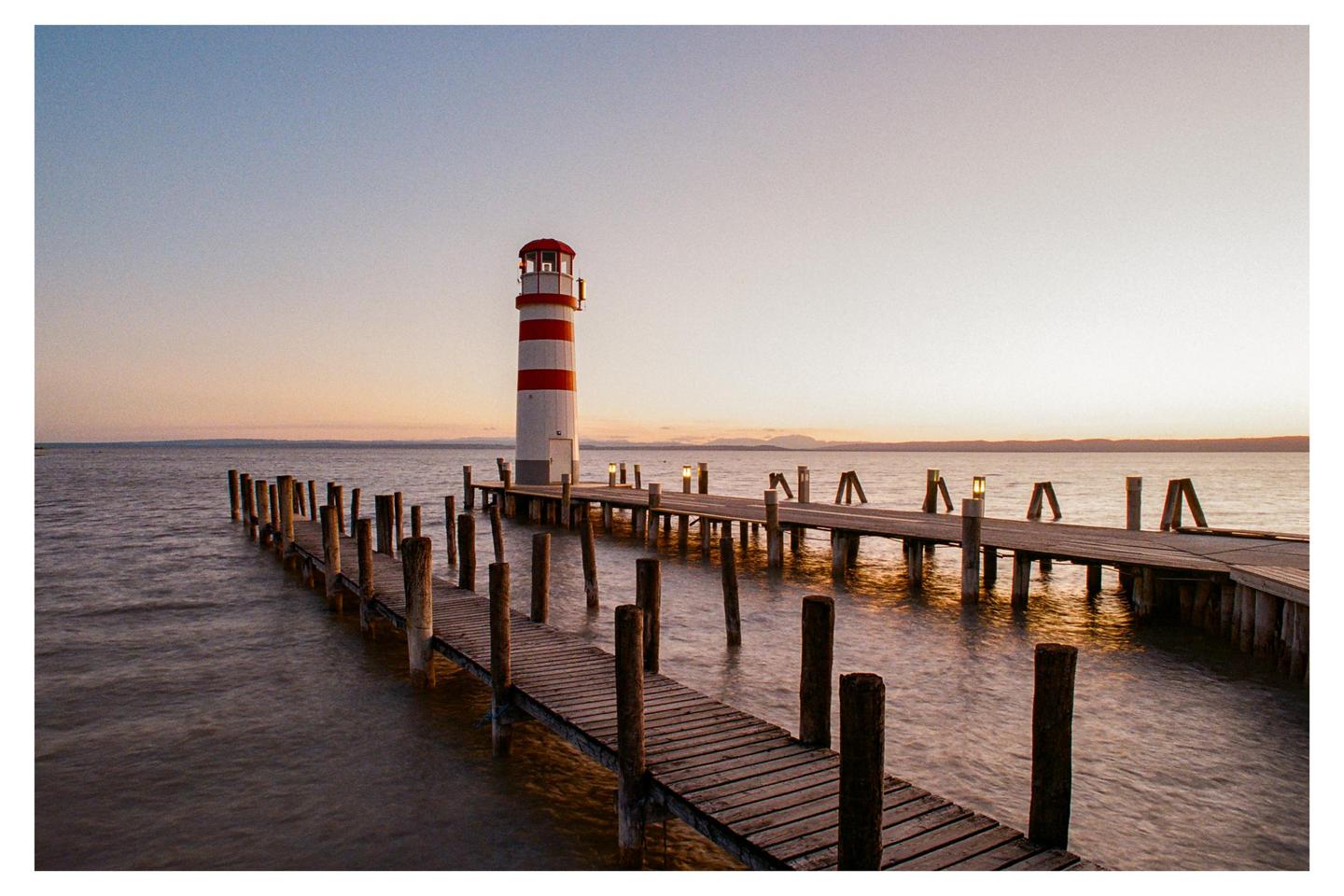 A red and white striped lighthouse stands at the end of a wooden pier on a lake at sunset.