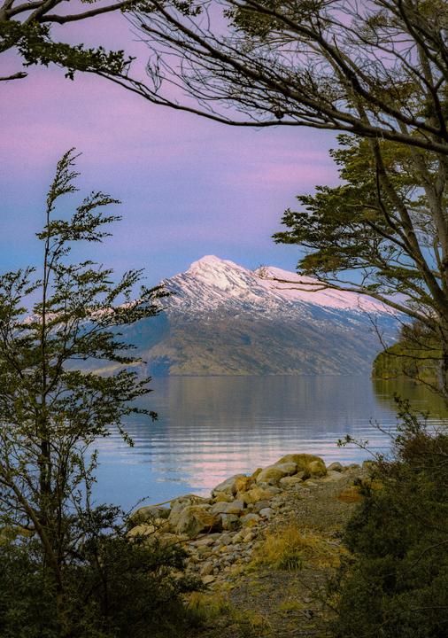 A snow-capped mountain rises above a lake, framed by trees and a rocky shore.