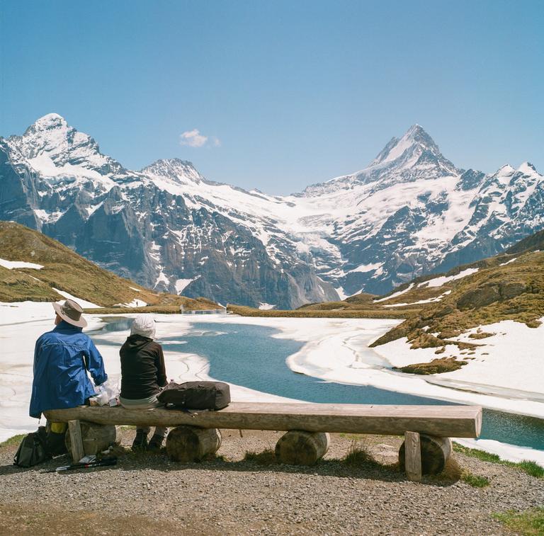 Two people sit on a wooden bench overlooking a partially frozen lake with snow-capped mountains in the background.