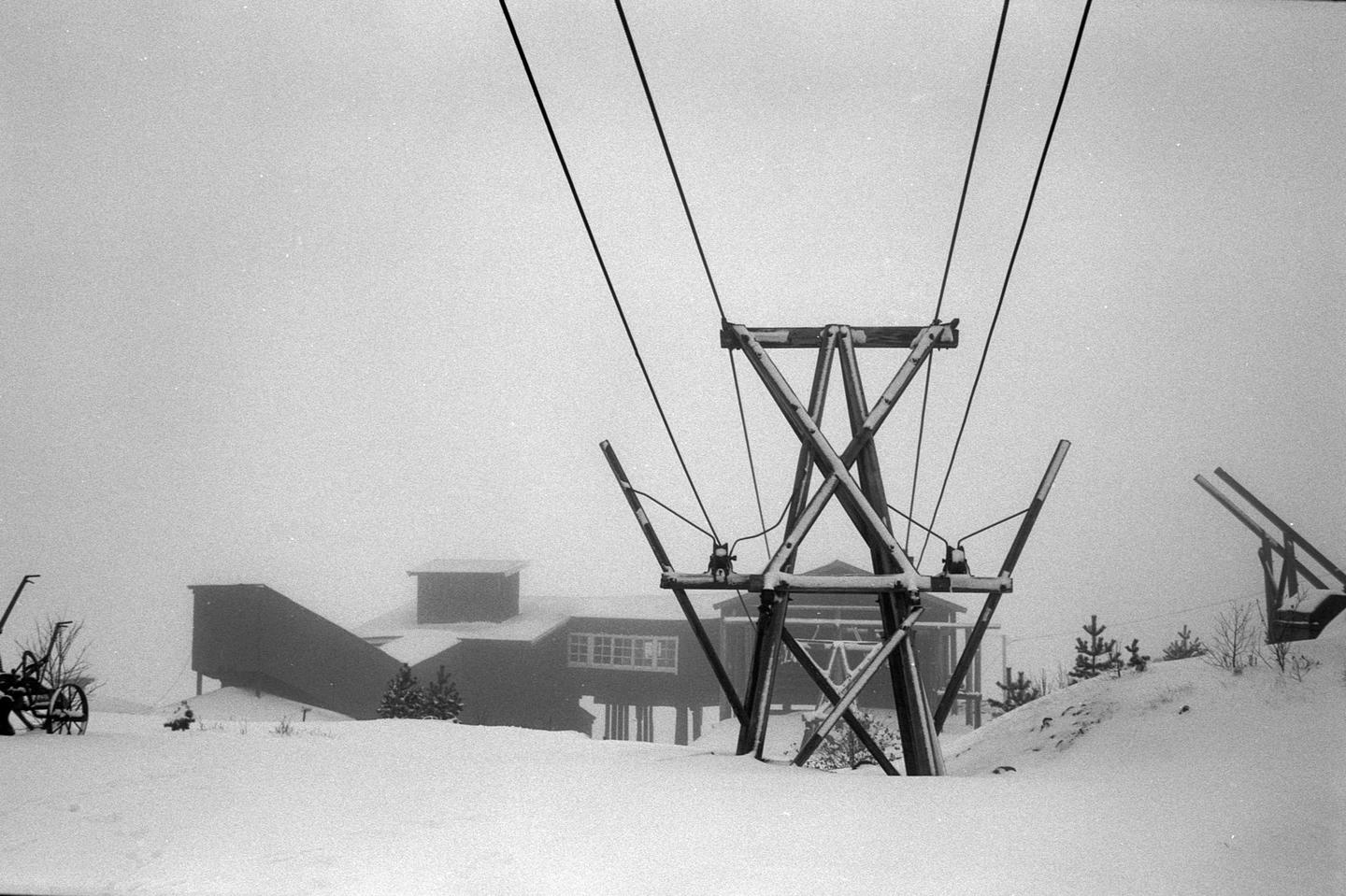 A ski lift tower stands in a snow-covered landscape with a building in the background.