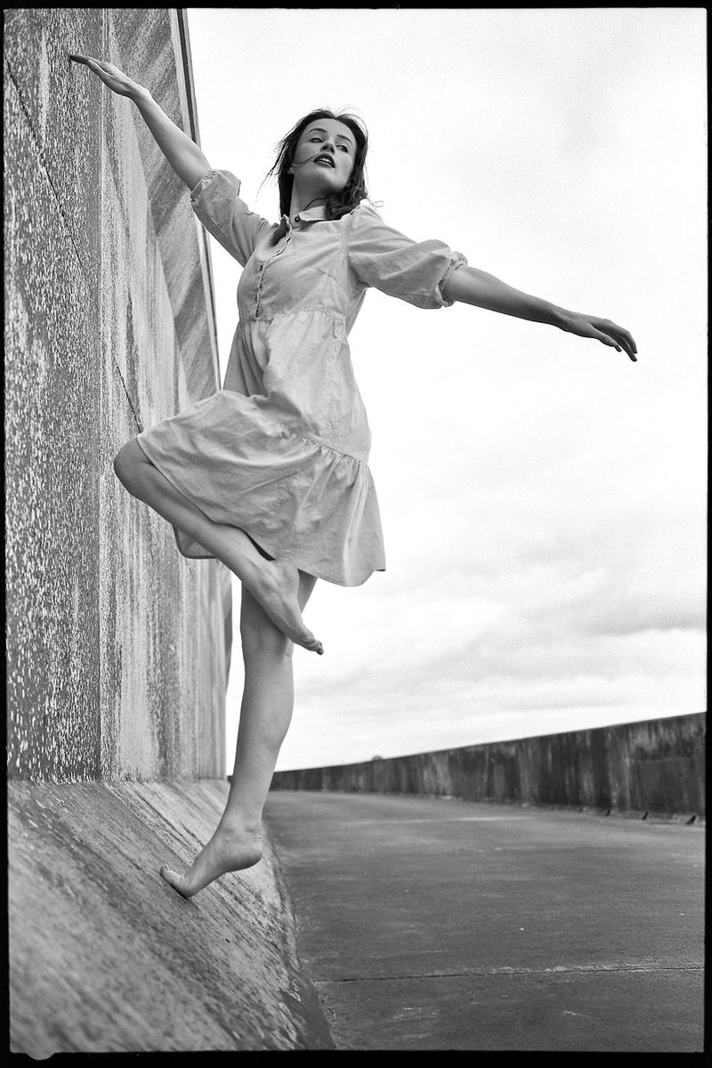 A woman in a dress balances on a concrete ledge with her arms outstretched.