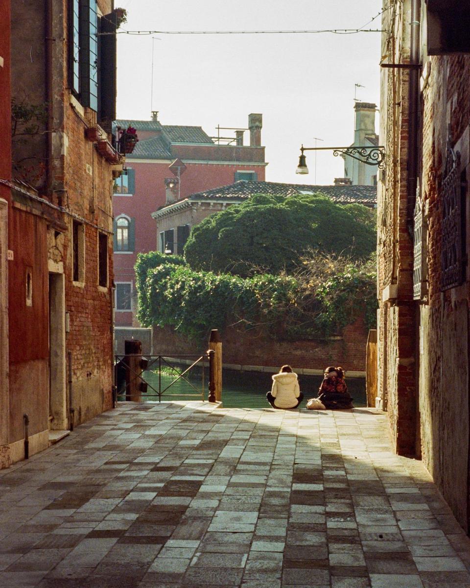 Two people sit on a stone path next to a canal in Venice, surrounded by brick buildings and lush greenery.