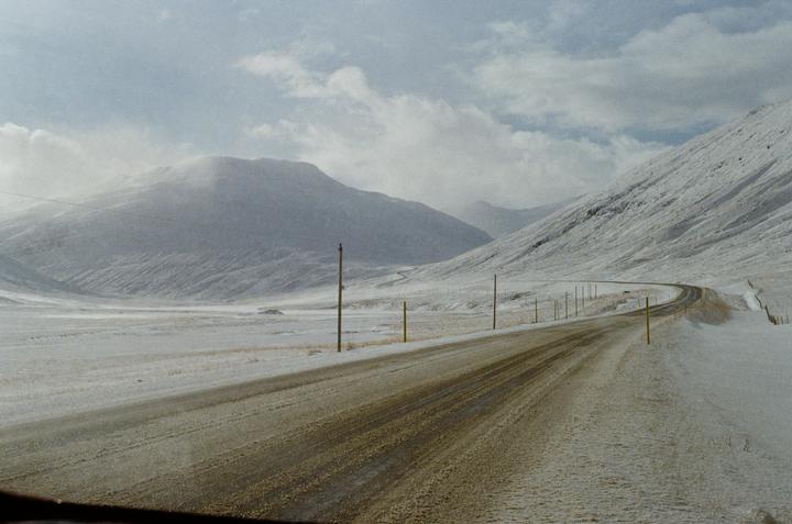A winding road cuts through a snow-covered valley with mountains under a cloudy sky.