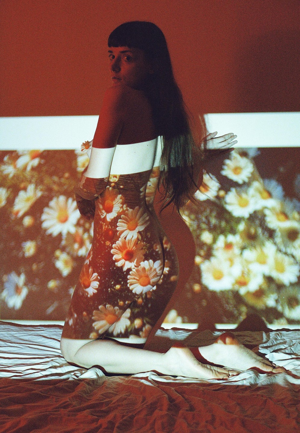 A woman kneels on a bed with a floral pattern projected onto her body and the wall behind her.