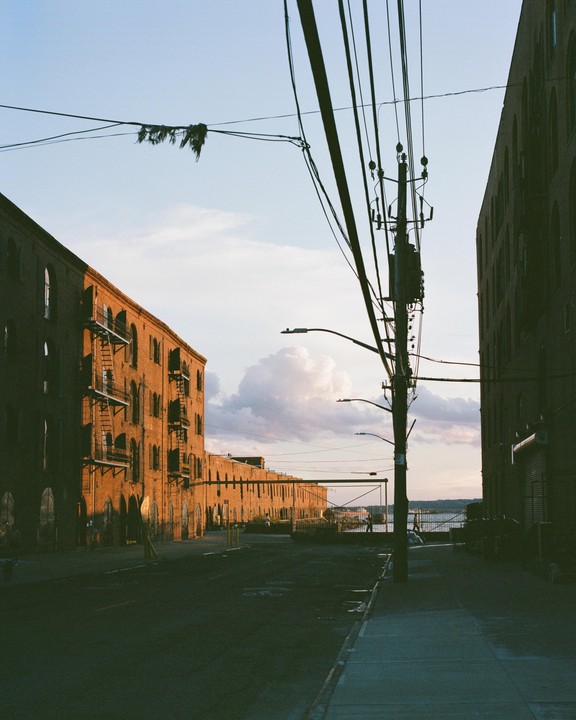 A street with brick buildings on either side leads to a body of water under a cloudy sky.