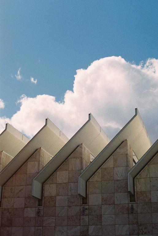 The sawtooth roof of a building with a tiled facade is visible against a blue sky with white clouds.