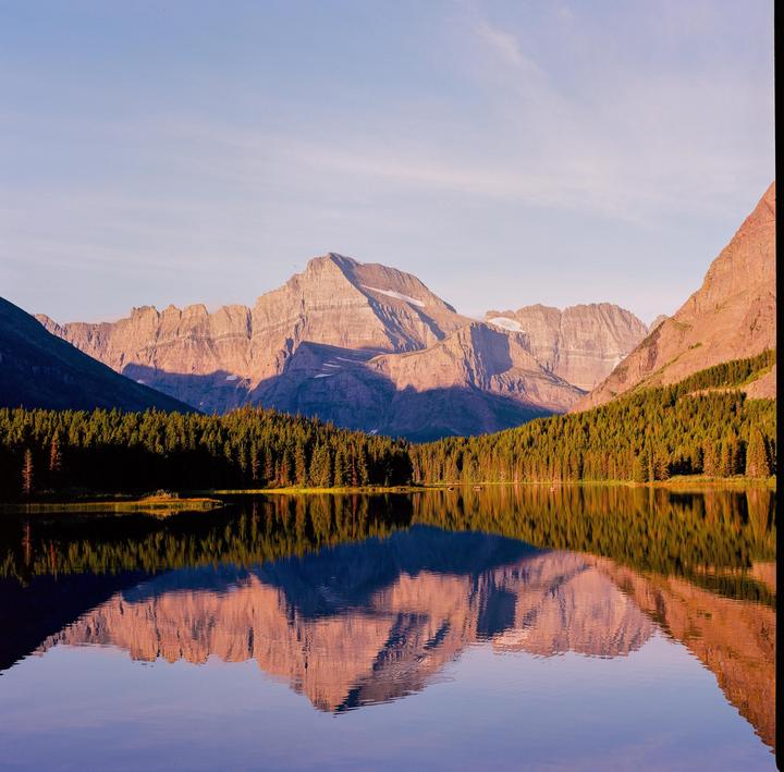A mountain range with snow-capped peaks is reflected in a calm lake surrounded by a dense forest.