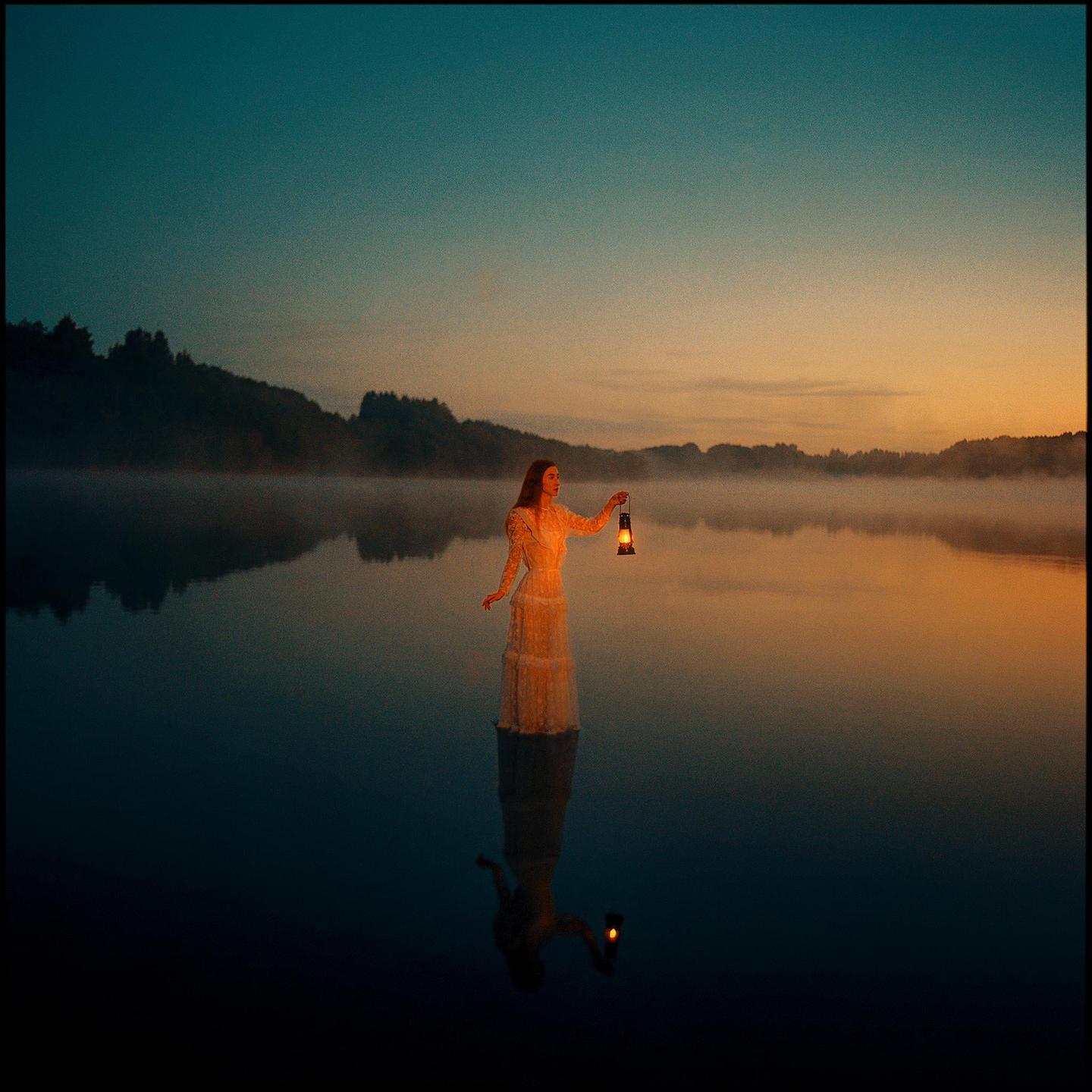 A woman in a long dress stands in a misty lake holding a lit lantern at dusk.