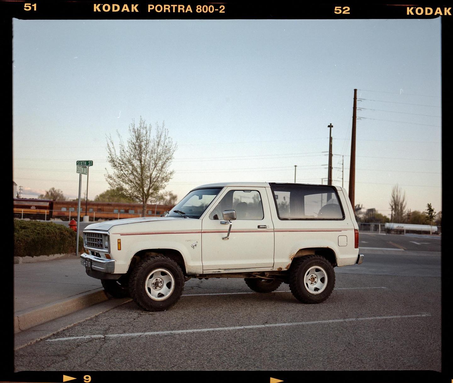 A white Ford Bronco is parked on a street next to a curb with a street sign in the background.