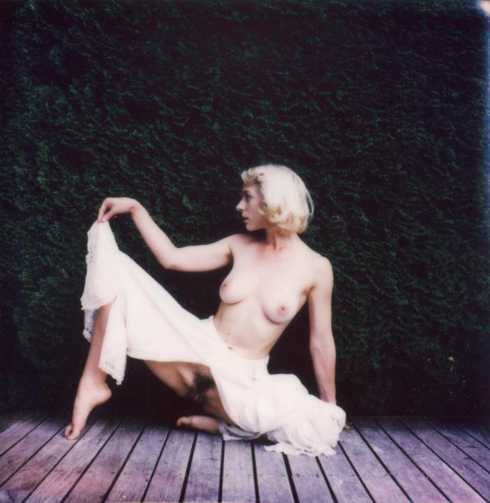A woman with blonde hair kneels on a wooden deck, holding up a white garment, with a dark green hedge in the background.