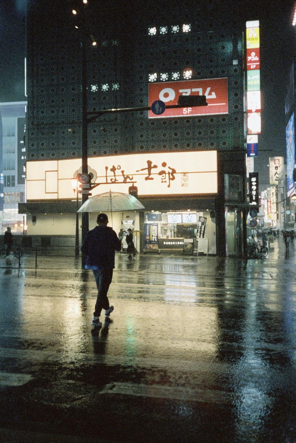 A man with an umbrella walks down a wet street at night, with neon signs and buildings in the background.