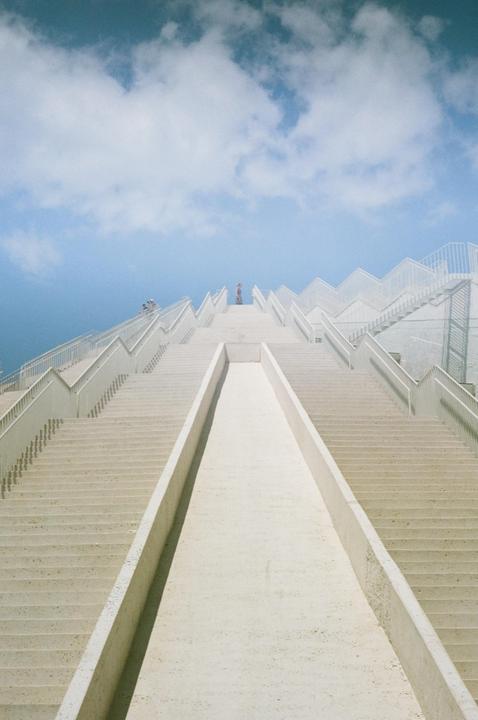 A person stands at the top of a wide, white, zig-zagging staircase under a blue sky with clouds.