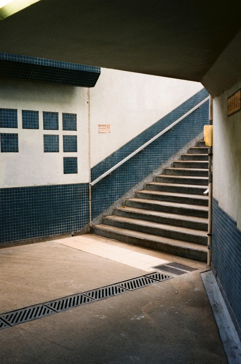 A concrete staircase with a blue tiled wall and handrail leads upwards from a tiled ground with a drain.
