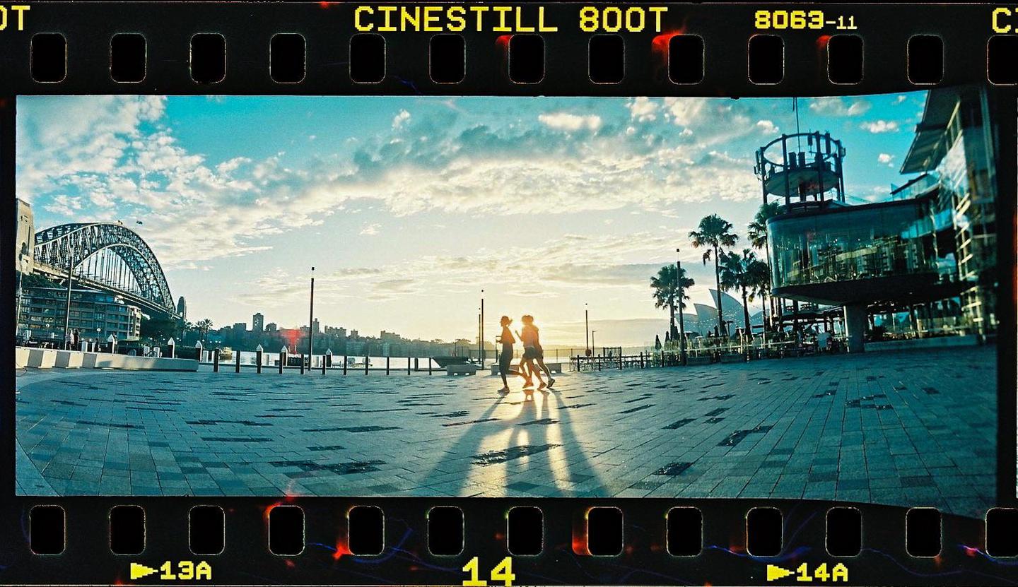 Three people run across a paved area with a bridge and modern buildings in the background under a cloudy sky.