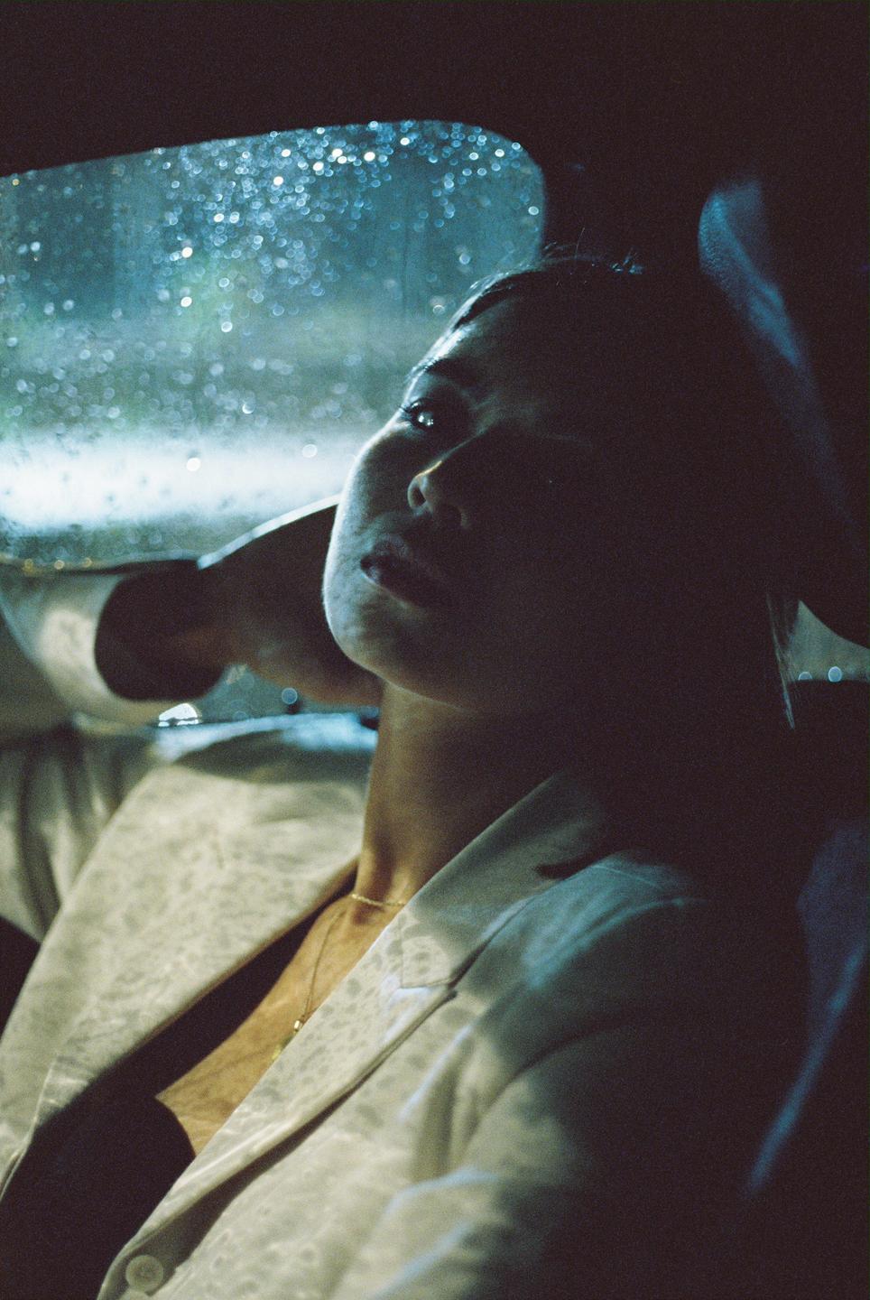 A woman sits in a car at night, looking up at the rain-streaked window.