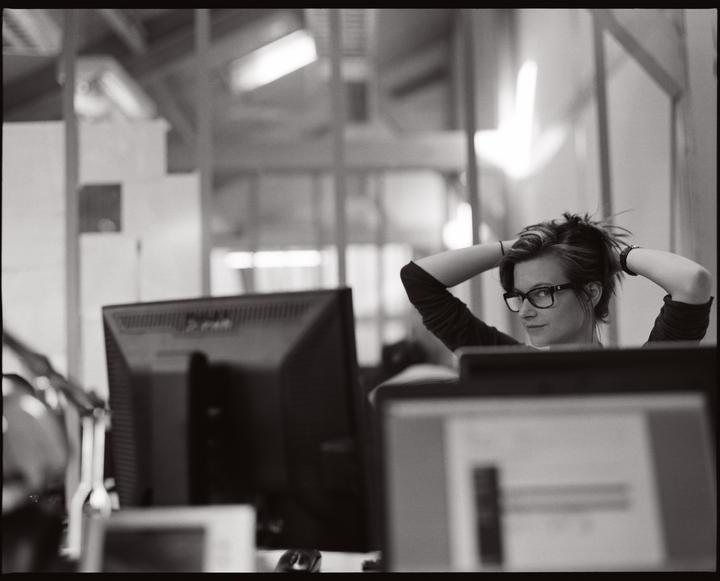 A woman with glasses is sitting at a desk with her hands behind her head, looking over her shoulder at the viewer.