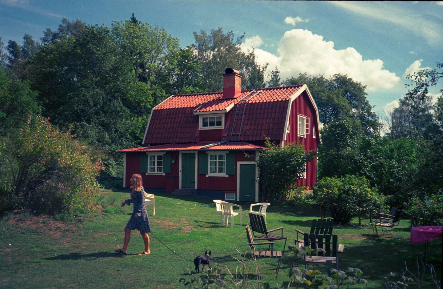 A woman walks a small dog on a leash in a grassy yard in front of a red house with a red roof and green shutters.