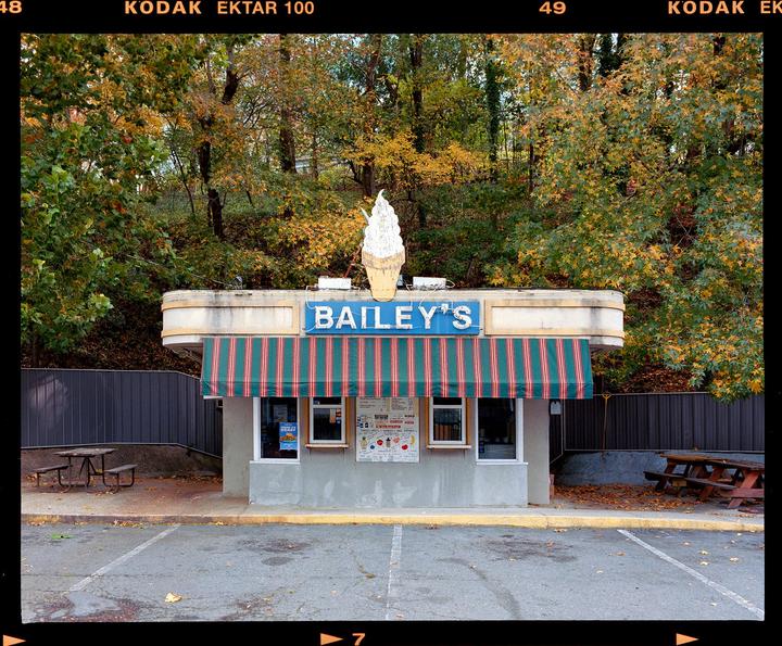 An ice cream stand with a large ice cream cone sign on its roof is surrounded by trees with autumn foliage.