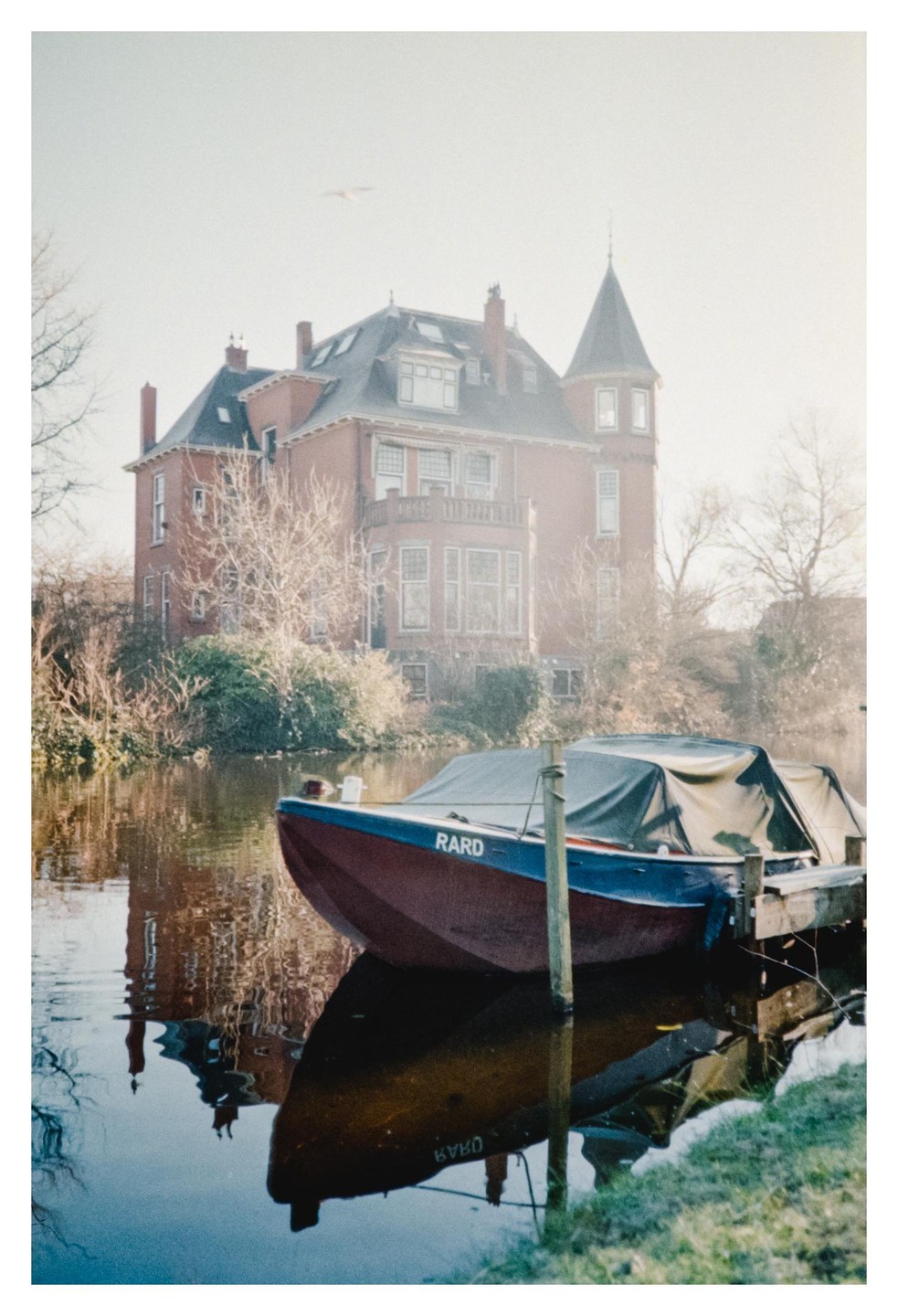 A boat is docked on a canal with a large brick house in the background on a misty morning.
