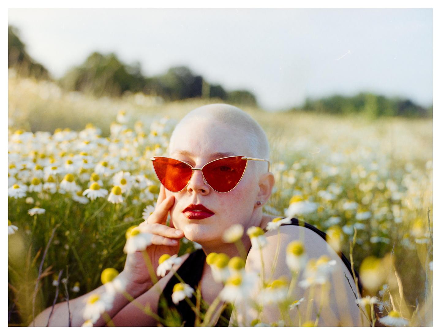 A woman with a shaved head and red sunglasses is posing in a field of white and yellow flowers.
