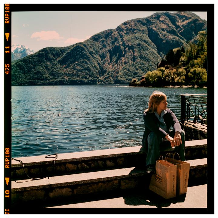 A woman sits on stone steps by a lake with mountains in the background.