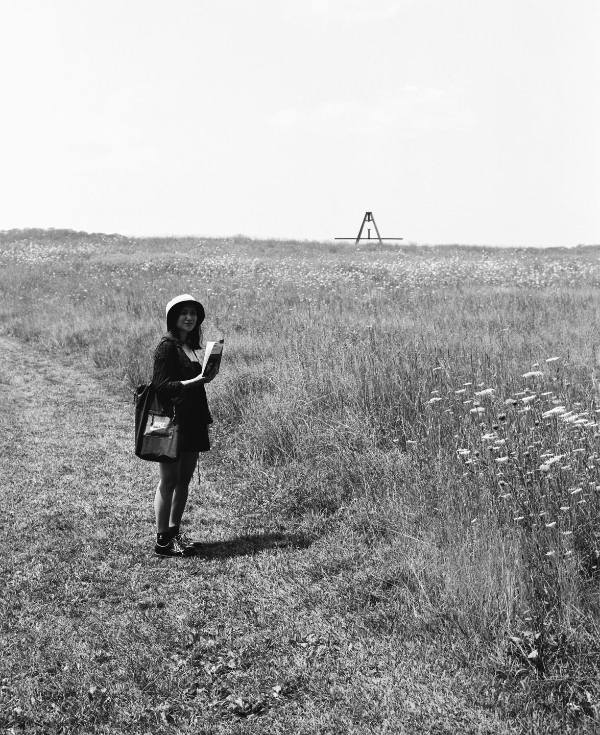 A woman wearing a hat and carrying a bag stands in a field of tall grass and wildflowers, holding a book.