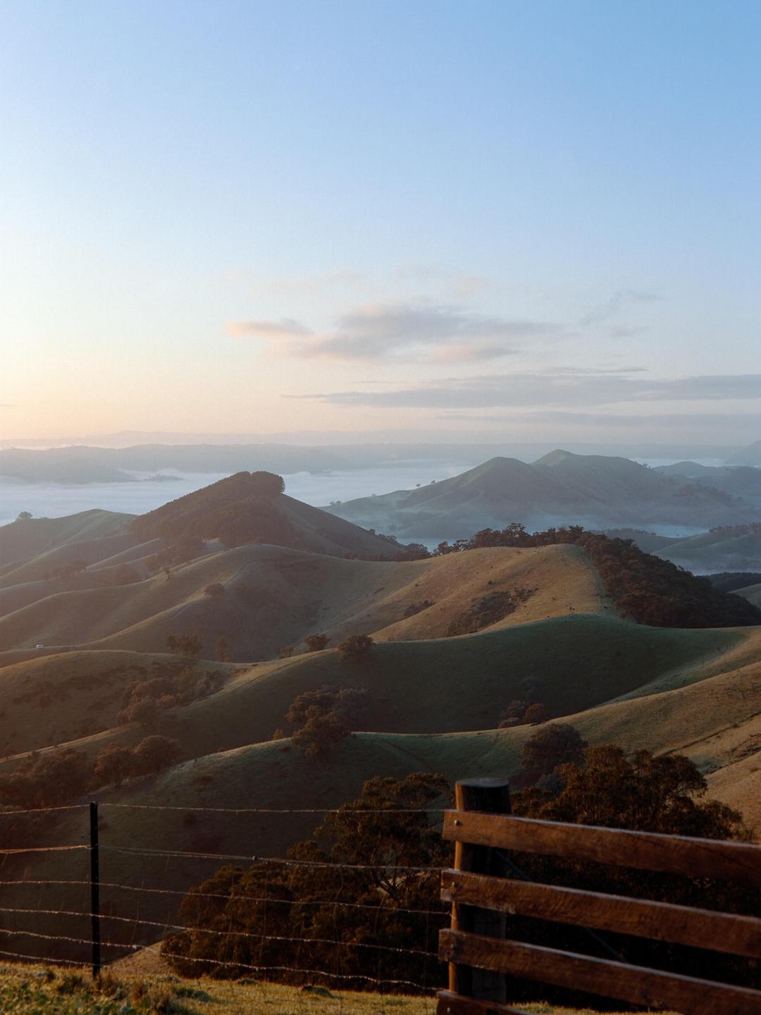 A wooden fence and barbed wire fence are in the foreground of rolling hills covered in fog.
