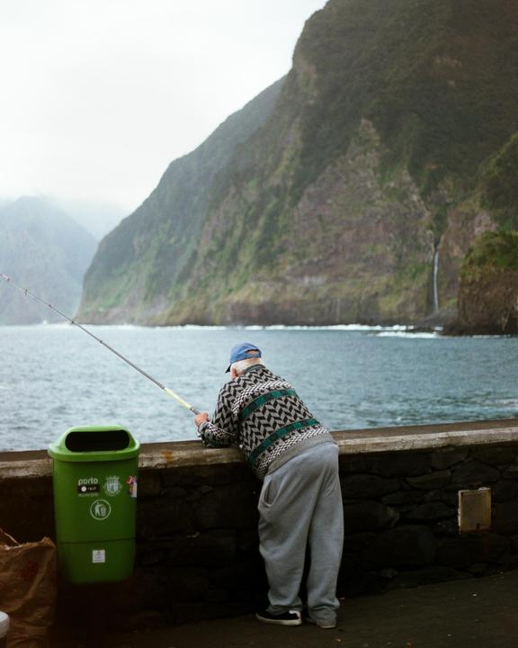 A man in a patterned sweater and blue cap fishes from a stone wall overlooking the ocean with a mountain and waterfall in the background.