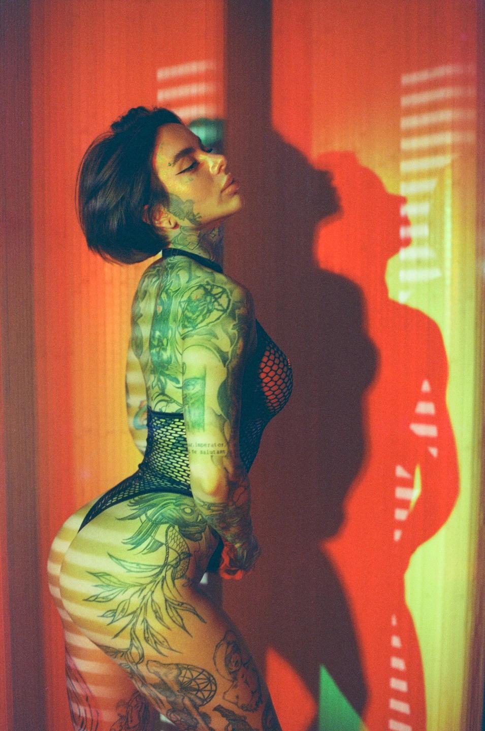 A woman with extensive tattoos poses in a fishnet bodysuit against a red wall with a strong shadow.