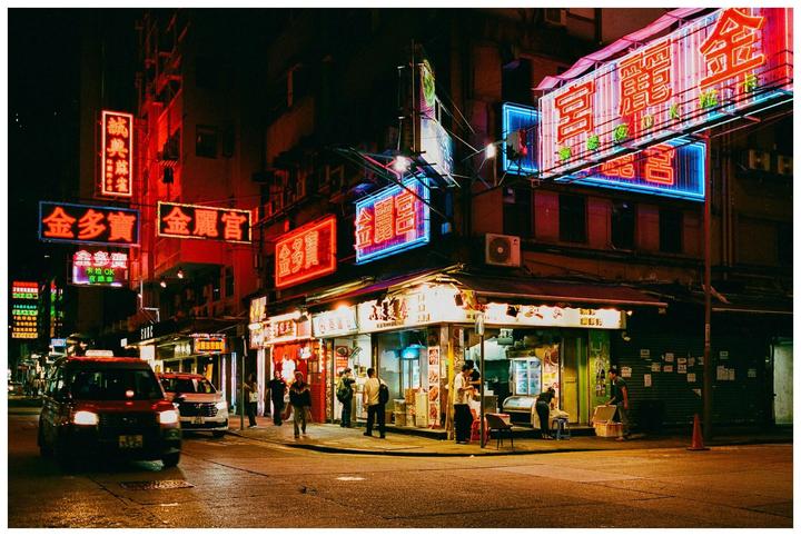 A street corner in Hong Kong is illuminated by numerous neon signs at night, with people and vehicles on the street.