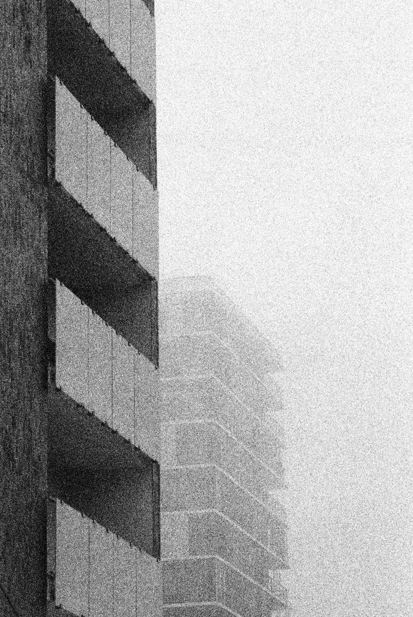 A tall building with balconies is partially obscured by fog, with another building faintly visible in the background.