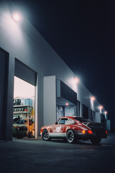 A red Porsche is parked outside a garage with its door open, revealing another vehicle and shelves of items inside.