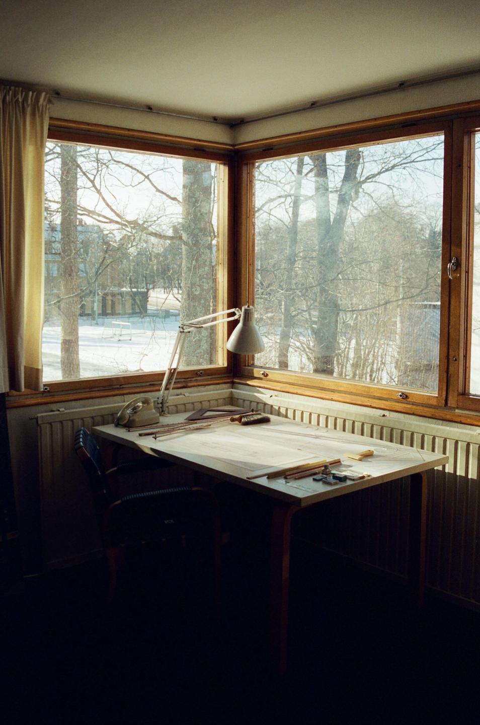 A drafting table with a lamp and tools sits in a corner by a large window overlooking a snowy landscape.