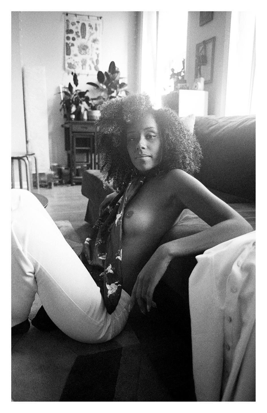 A woman with curly hair sits on the floor in a living room, wearing white pants and a scarf.