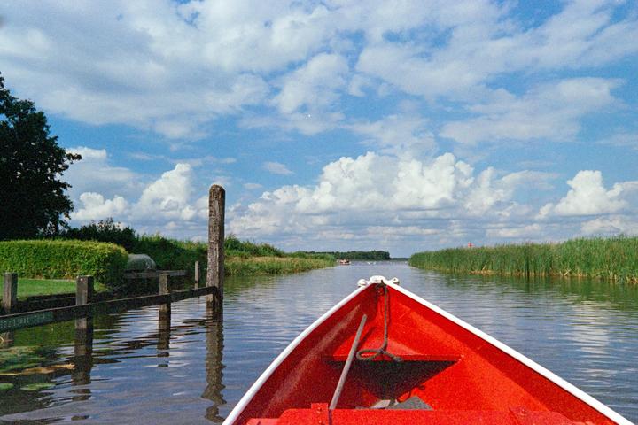 A red boat floats on a canal with green reeds on either side under a blue sky with white clouds.