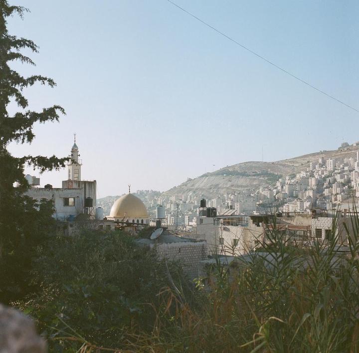 A mosque with a golden dome and a minaret stands among buildings on a hillside, with more buildings covering a distant mountain.