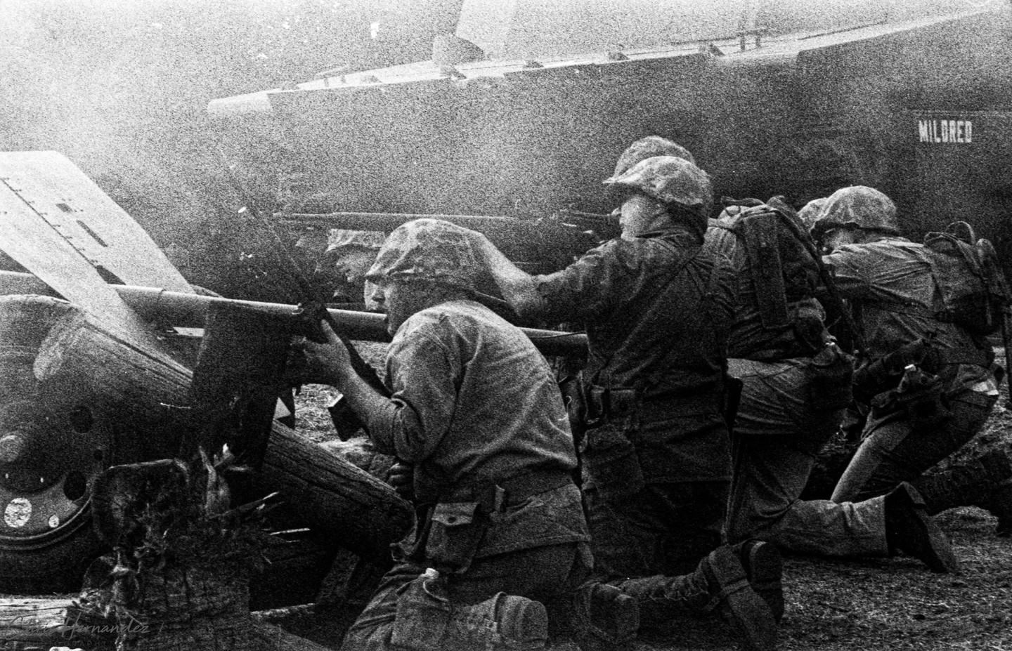 Several soldiers in helmets and uniform kneel with rifles in a battle reenactment, with a vehicle named "MILDRED" in the background.