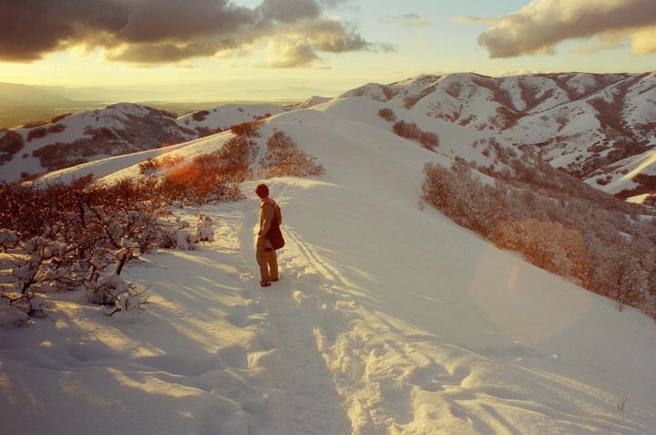 A person stands on a snowy mountain path looking out at a valley during golden hour.