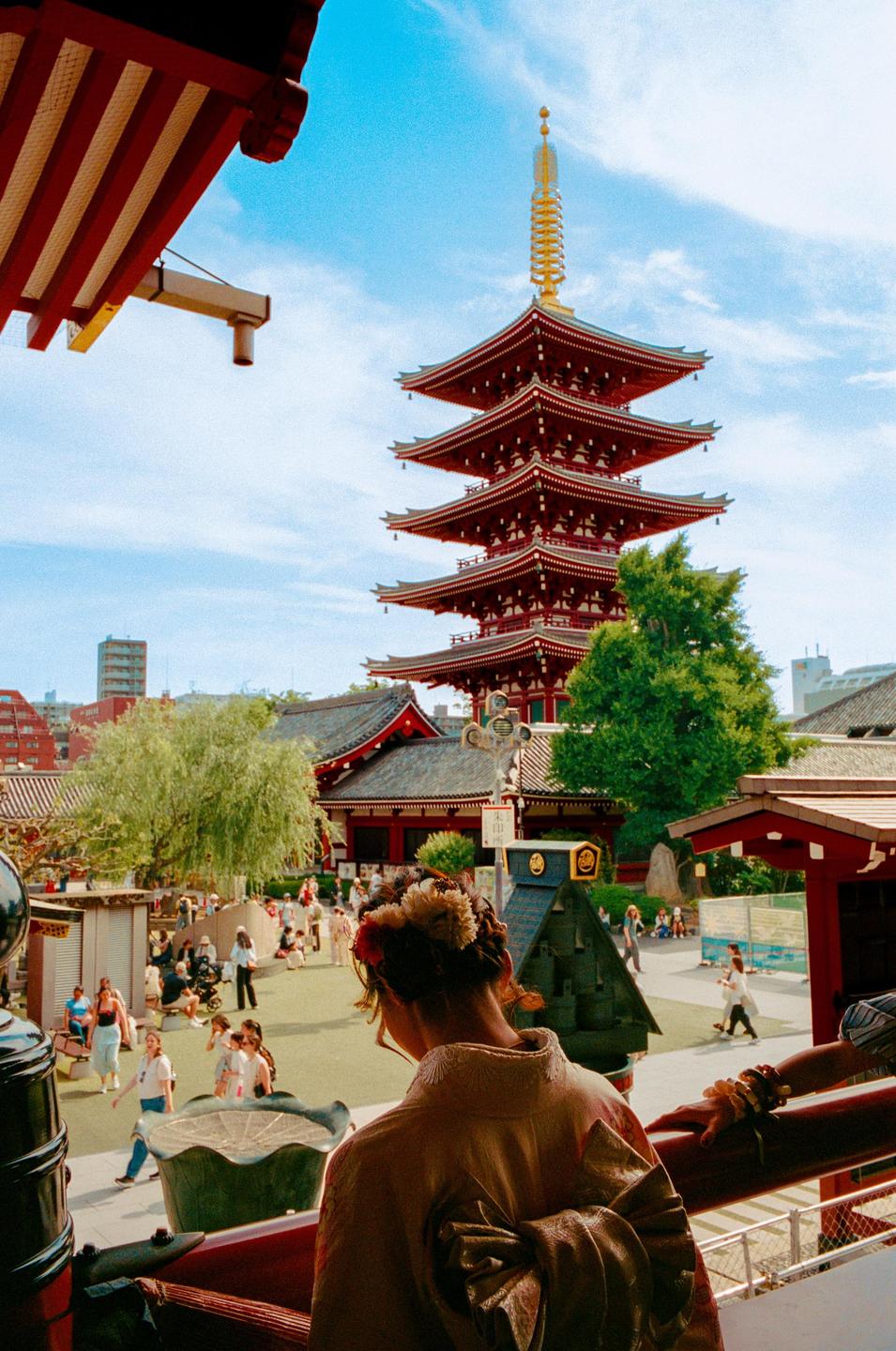 A woman in traditional dress looks out over a temple courtyard with a red pagoda in the background.