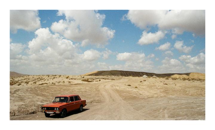 An orange car is parked on a dirt road in a desert landscape under a blue sky with clouds.