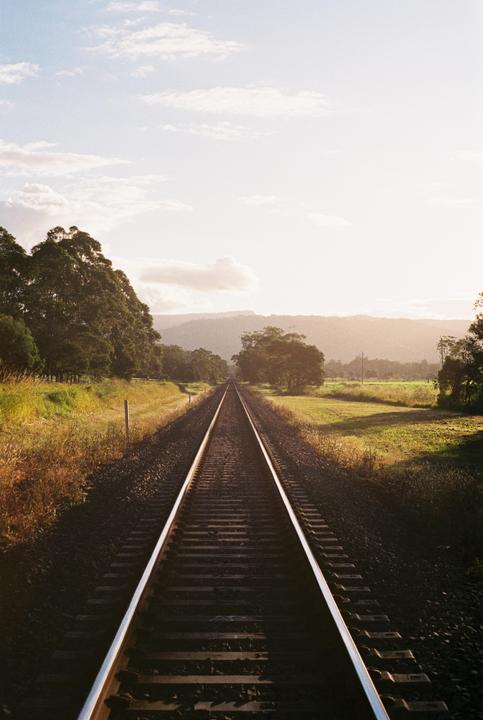 A long railway track stretches into the distance, flanked by trees and fields under a bright sky.